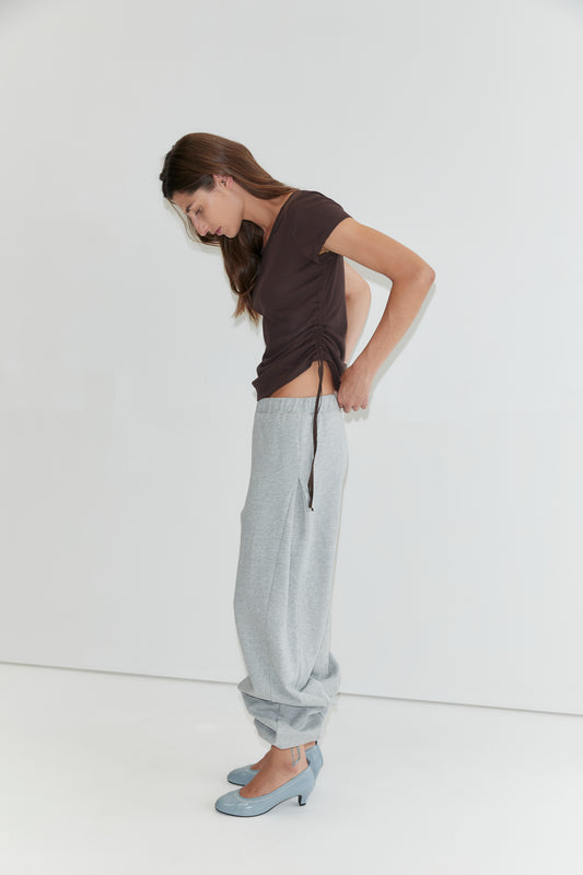 A woman in the Draw Side Tee - Brown by Deiji Studios and loose gray sweatpants, rolled up at the ankles, stands against a white background, looking down as she adjusts her cotton jersey shirt. She wears light blue high-heeled shoes.