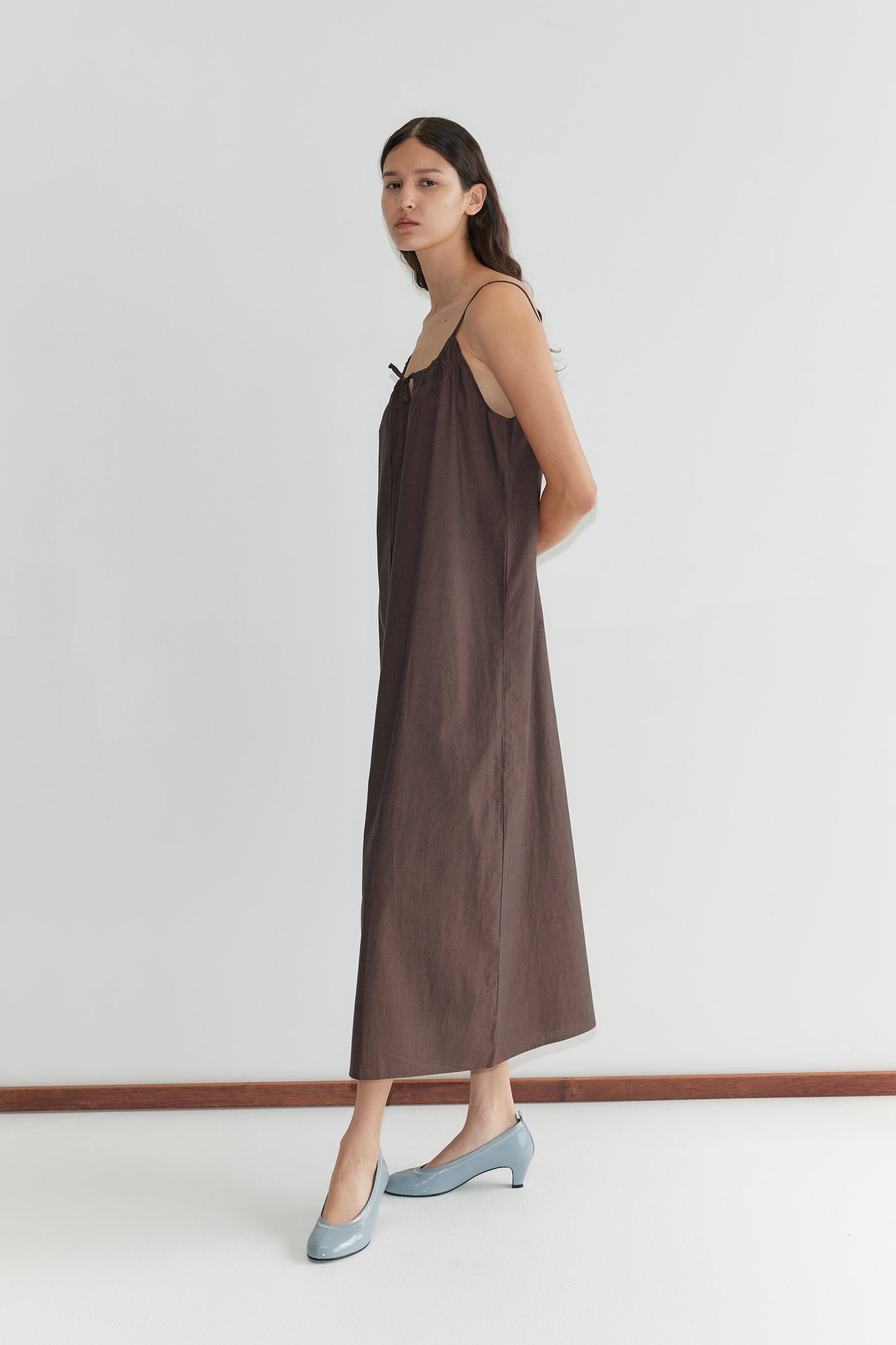 A woman stands in profile against a plain white wall, wearing the Light Maxi Dress - Toast from Deiji Studios with light blue shoes. Her long dark hair is down and she has a neutral expression.