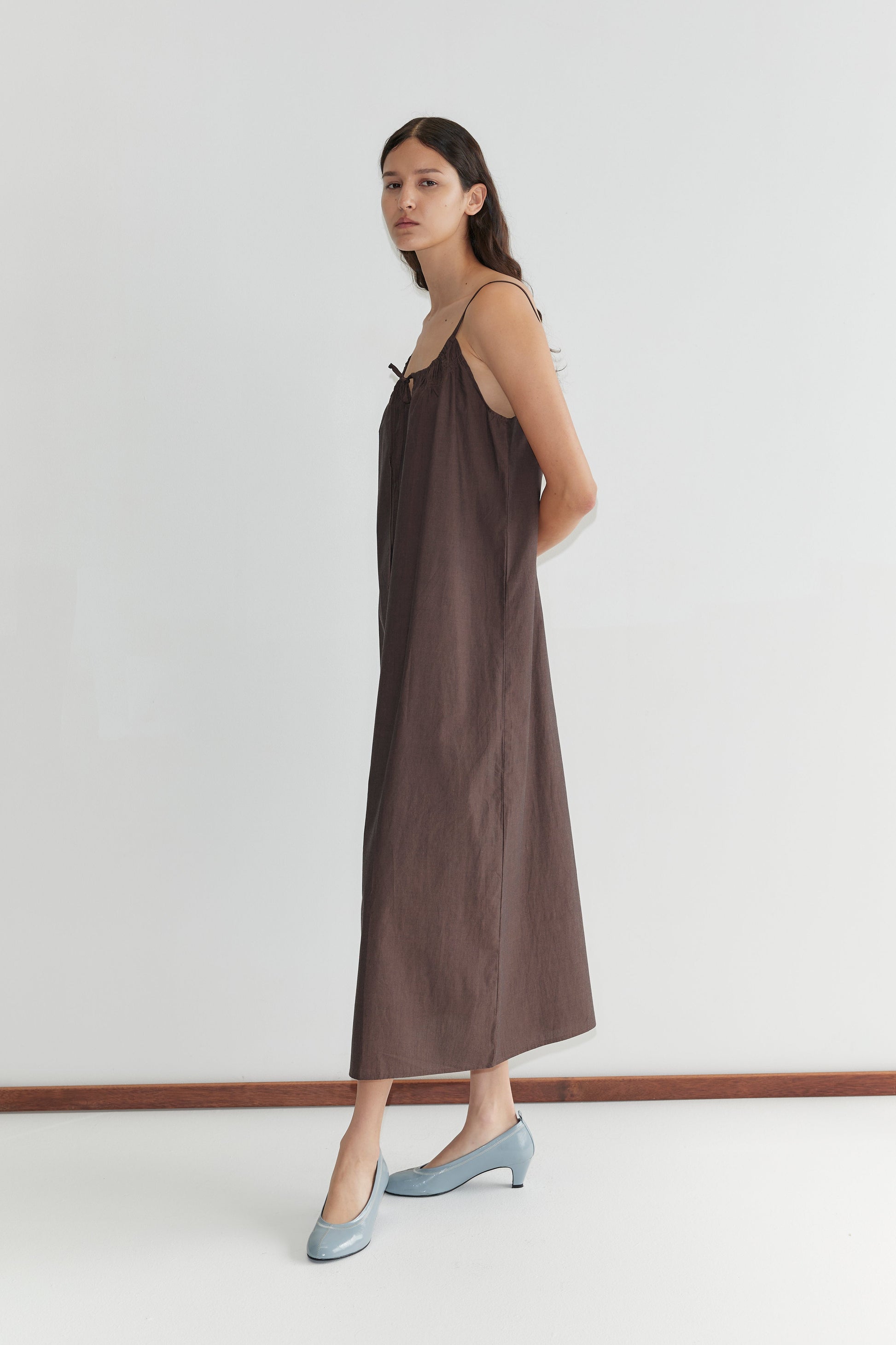 A woman stands in profile against a plain white wall, wearing the Light Maxi Dress - Toast from Deiji Studios with light blue shoes. Her long dark hair is down and she has a neutral expression.