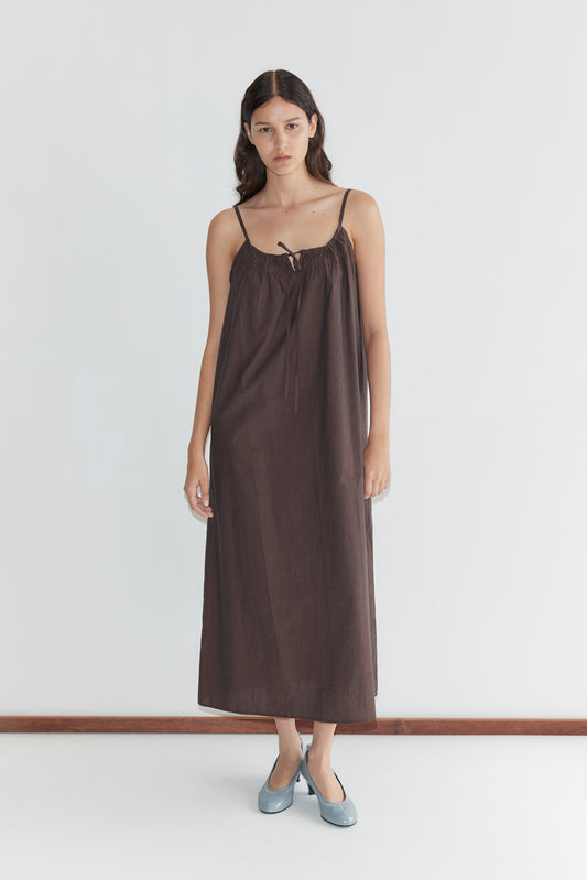 A woman with long, dark hair and a neutral expression stands against a plain white wall, wearing the Light Maxi Dress - Toast by Deiji Studios with thin straps and light blue flat shoes.