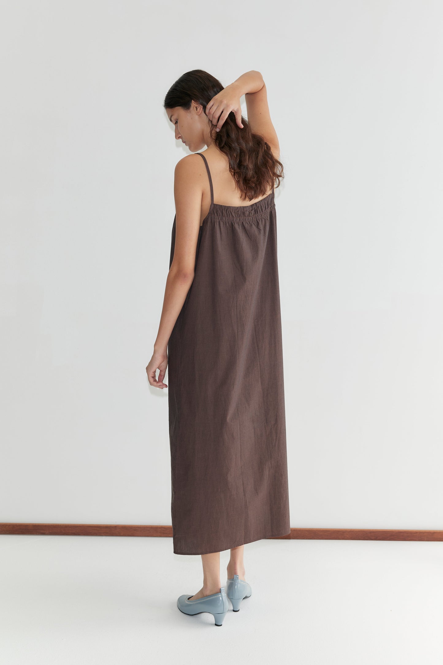 A woman in a relaxed Light Maxi Dress - Toast by Deiji Studios and light blue shoes stands against a white wall, facing away with one arm raised to touch the back of her head.