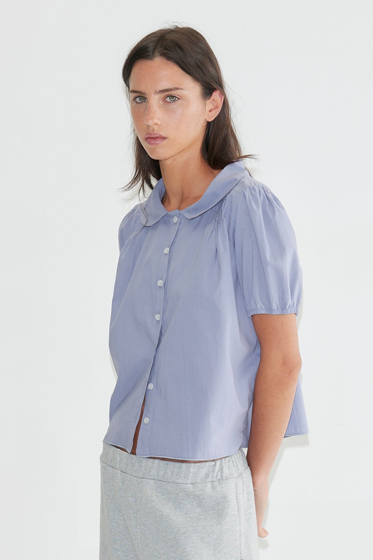 A woman with long dark hair stands against a plain white background wearing the Petal Top - Thistle by Deiji Studios, a pale periwinkle petal-collar blouse, with grey marle trousers and pale blue heels.