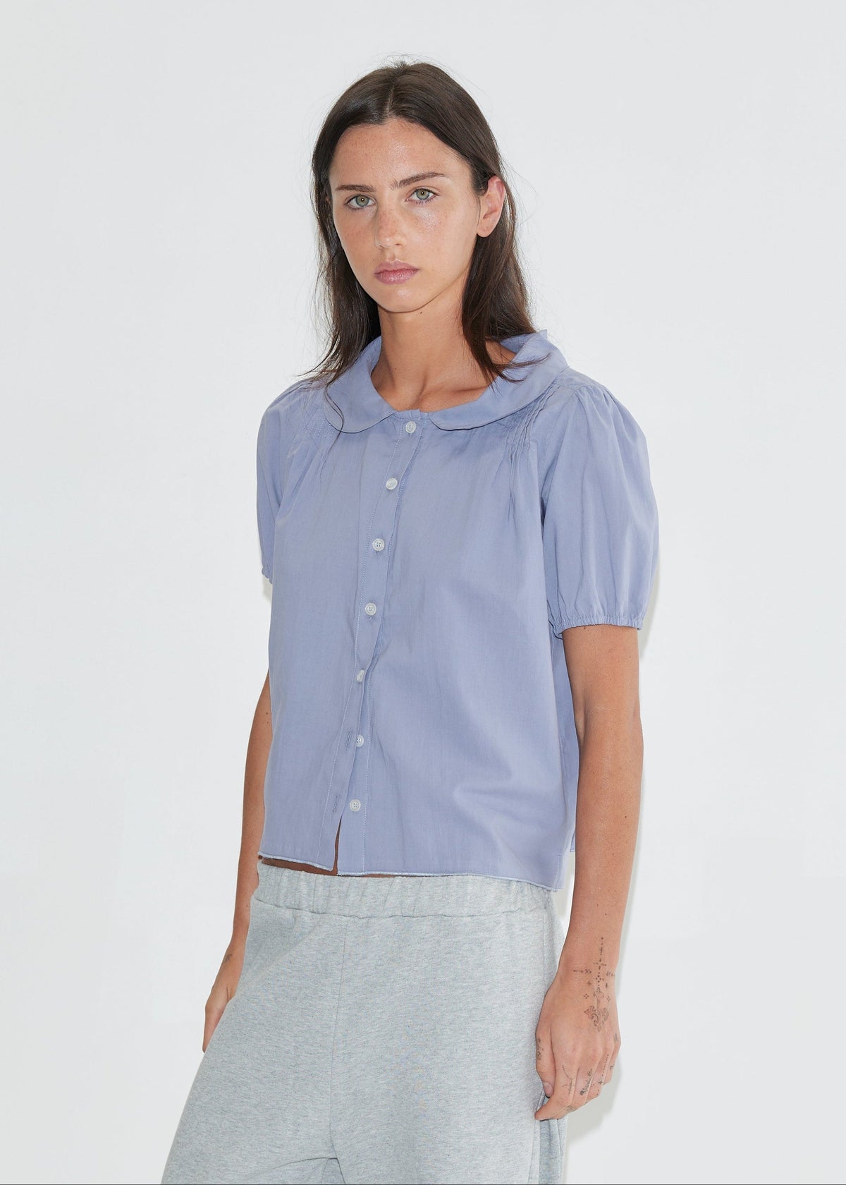 A cropped front view of the Petal Top - Thistle by Deiji Studios, a pale periwinkle short-sleeve blouse with a rounded petal collar, front buttons and puff sleeves, with grey marle trousers against a plain white background.