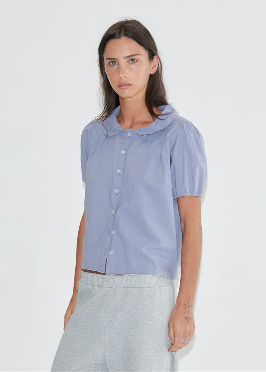 A cropped front view of the Petal Top - Thistle by Deiji Studios, a pale periwinkle short-sleeve blouse with a rounded petal collar, front buttons and puff sleeves, with grey marle trousers against a plain white background.