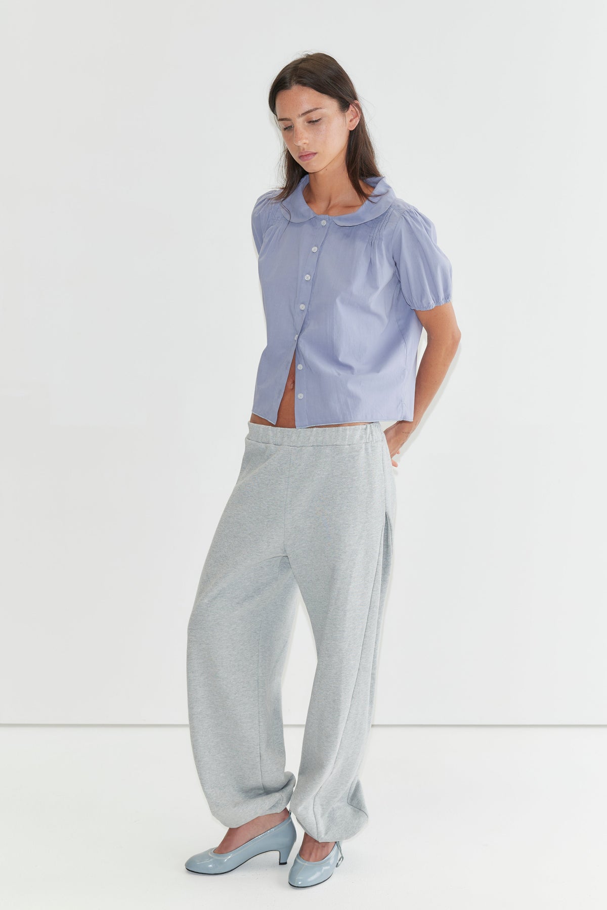 A woman with long dark hair stands against a plain white background looking down, wearing the Petal Top - Thistle by Deiji Studios, a pale periwinkle petal-collar blouse with puff sleeves, and grey marle trousers with pale blue heels.
