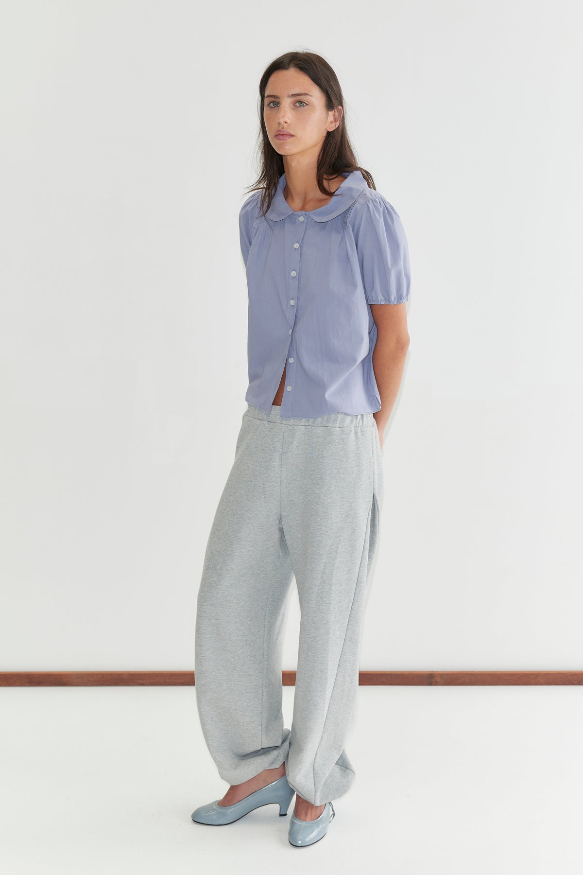 A woman with long dark hair stands against a plain white background wearing the Petal Top - Thistle by Deiji Studios, a pale periwinkle petal-collar blouse with front buttons and puff sleeves, with grey marle trousers and pale blue heels.