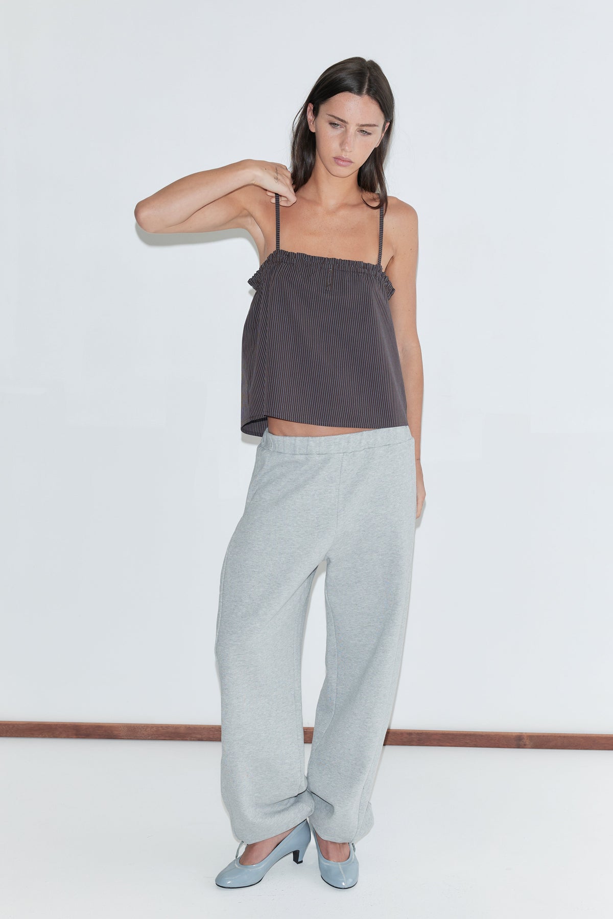 A woman with long dark hair stands against a plain white background wearing the Frill Placket Top - Brown Blue Stripe by Deiji Studios, a dark brown fine-striped cami top with thin straps and a gathered frill placket, with grey marle trousers and pale blue heels.