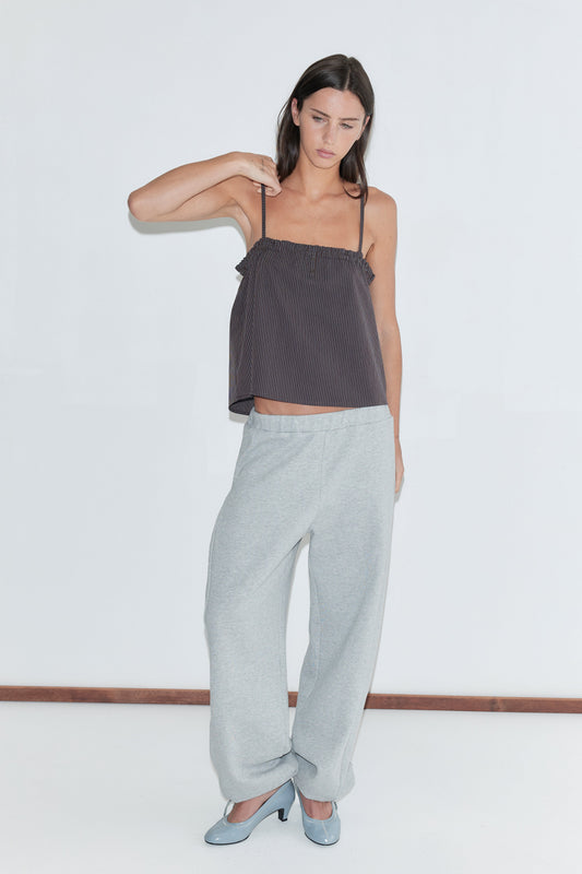 A woman with long dark hair stands against a plain white background wearing the Frill Placket Top - Brown Blue Stripe by Deiji Studios, a dark brown fine-striped cami top with thin straps and a gathered frill placket, with grey marle trousers and pale blue heels.