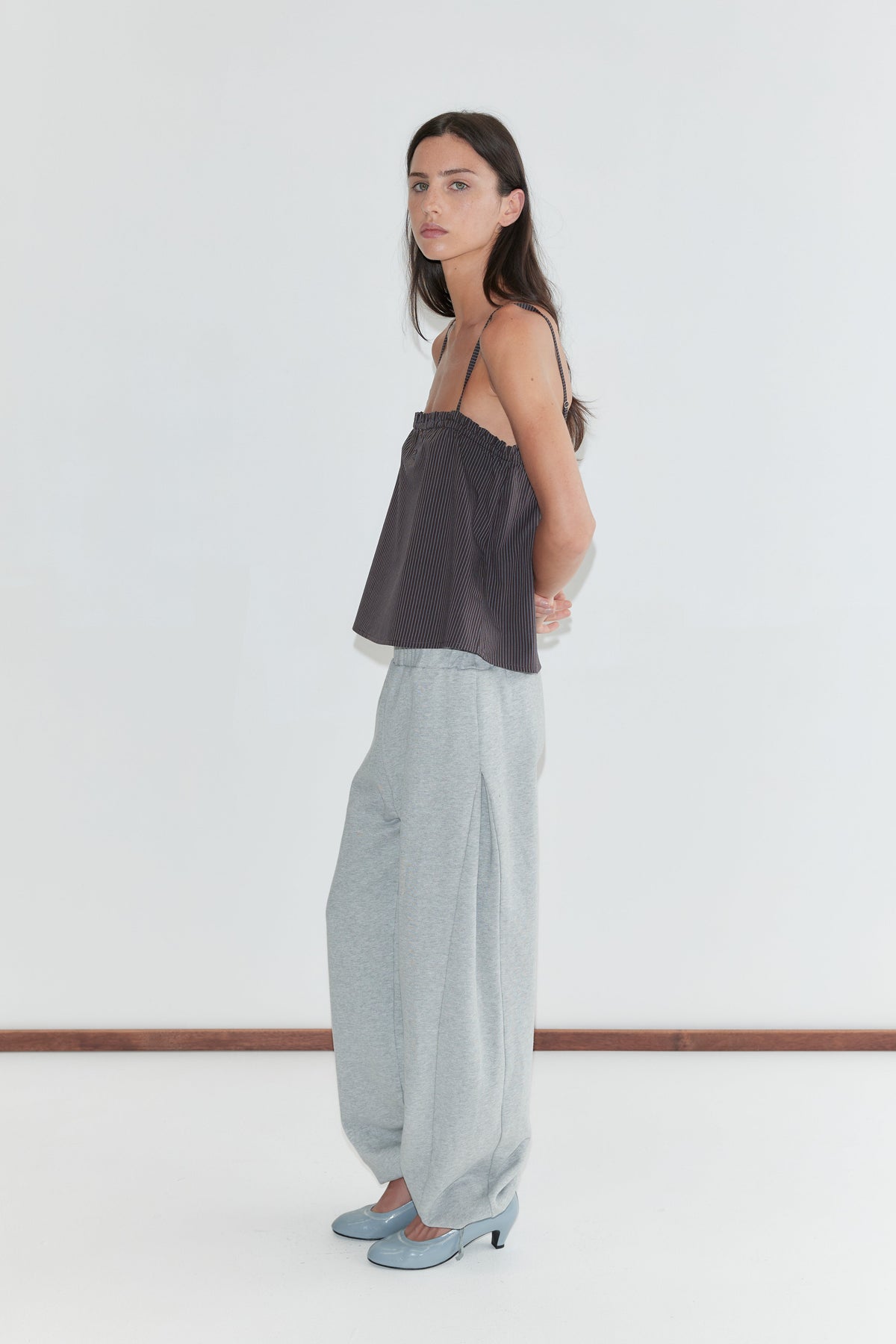 A woman with long dark hair stands in profile against a plain white background wearing the Frill Placket Top - Brown Blue Stripe by Deiji Studios, a dark brown fine-striped cami top, with grey marle trousers and pale blue heels.