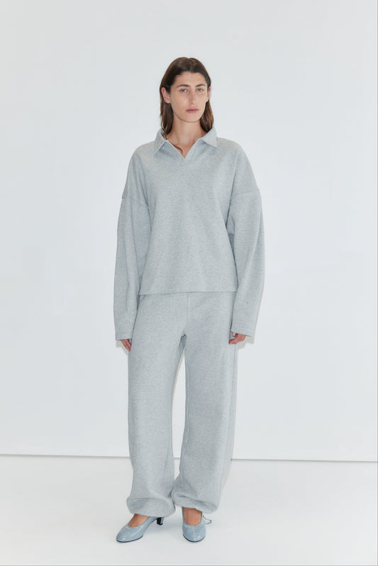 A person stands against a white background, wearing a loose light gray sweater paired with Deiji Studios' Side Pleat Pant in Grey Marle and light blue heels.