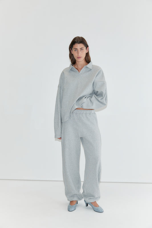 A person stands against a white background wearing the Jersey Jumper - Grey Marle, styled with matching sweatpants and blue heels, with one hand tucked into their pocket.