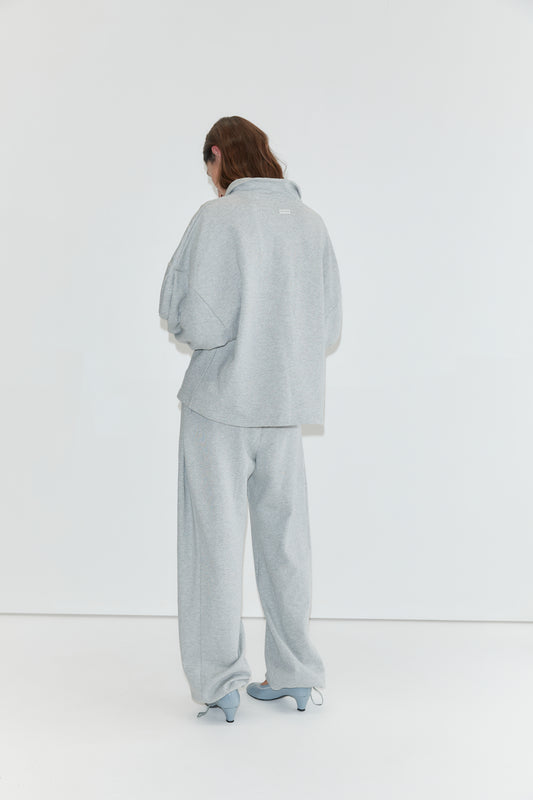A person stands facing a white wall, wearing the Jersey Jumper - Grey Marle, a loose light gray cotton jersey jumper with an open polo collar, paired with matching pants and light blue heeled shoes. Their long hair partially covers their face.