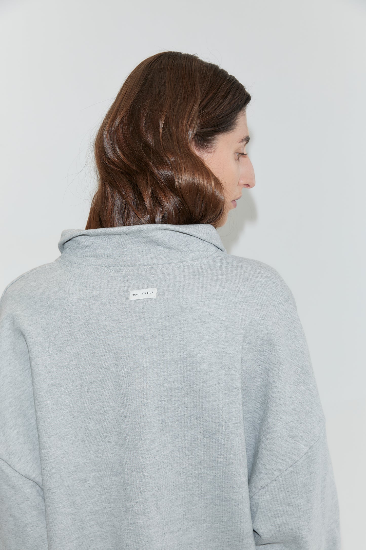 A person with long brown hair, facing away from the camera, wears the Jersey Jumper in Grey Marle. The light gray cotton sweatshirt features a small white label with text on the upper back. The background is plain and light-colored.
