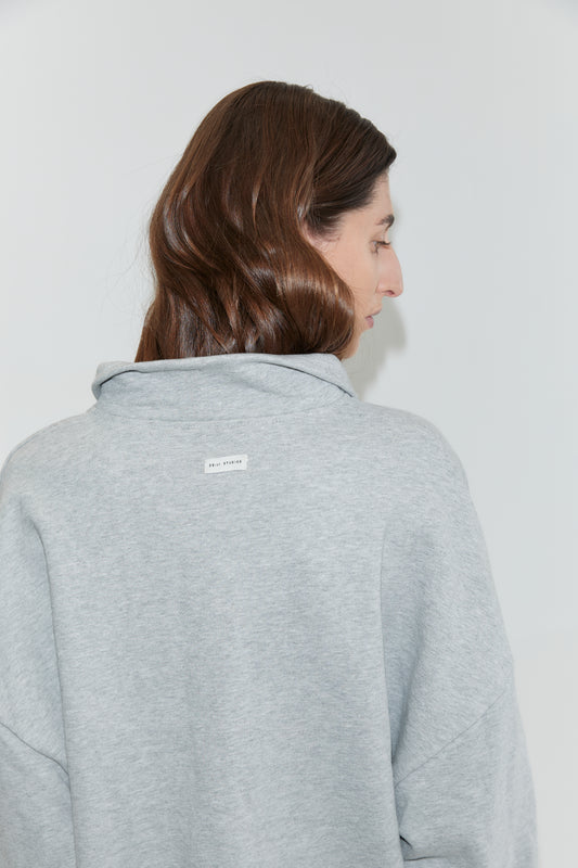 A person with long brown hair, facing away from the camera, wears the Jersey Jumper in Grey Marle. The light gray cotton sweatshirt features a small white label with text on the upper back. The background is plain and light-colored.