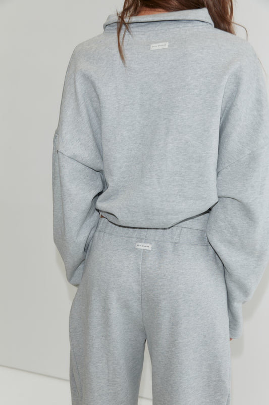A person wearing a matching light gray sweatshirt and wide-leg cotton pant set, shown from the back. The outfit, inspired by Deiji Studios, features white tags on the upper back of the top and on the waistband of the Side Pleat Pant - Grey Marle.
