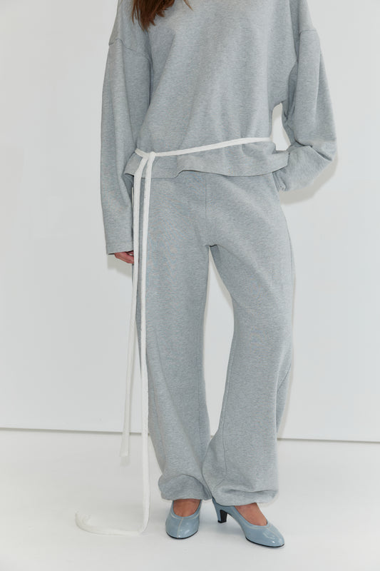 A person is shown wearing the Side Pleat Pant - Grey Marle, styled with a loose-fitting light gray sweatshirt and cinched at the waist with a long white belt, paired with blue high-heeled shoes. The face is not visible, capturing the relaxed elegance characteristic of Deiji Studios collections.