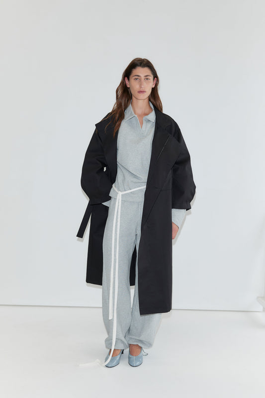 Wearing the Jersey Jumper in Grey Marle against a white background, the person completes their look with an open polo collar design, a white belt, a long black coat, and light blue shoes.