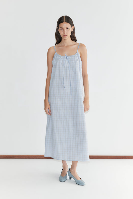 A woman with long dark hair wears the Light Maxi Dress - Powder Check, featuring thin straps and a gathered neckline. She pairs it with blue shoes and stands against a plain white background.