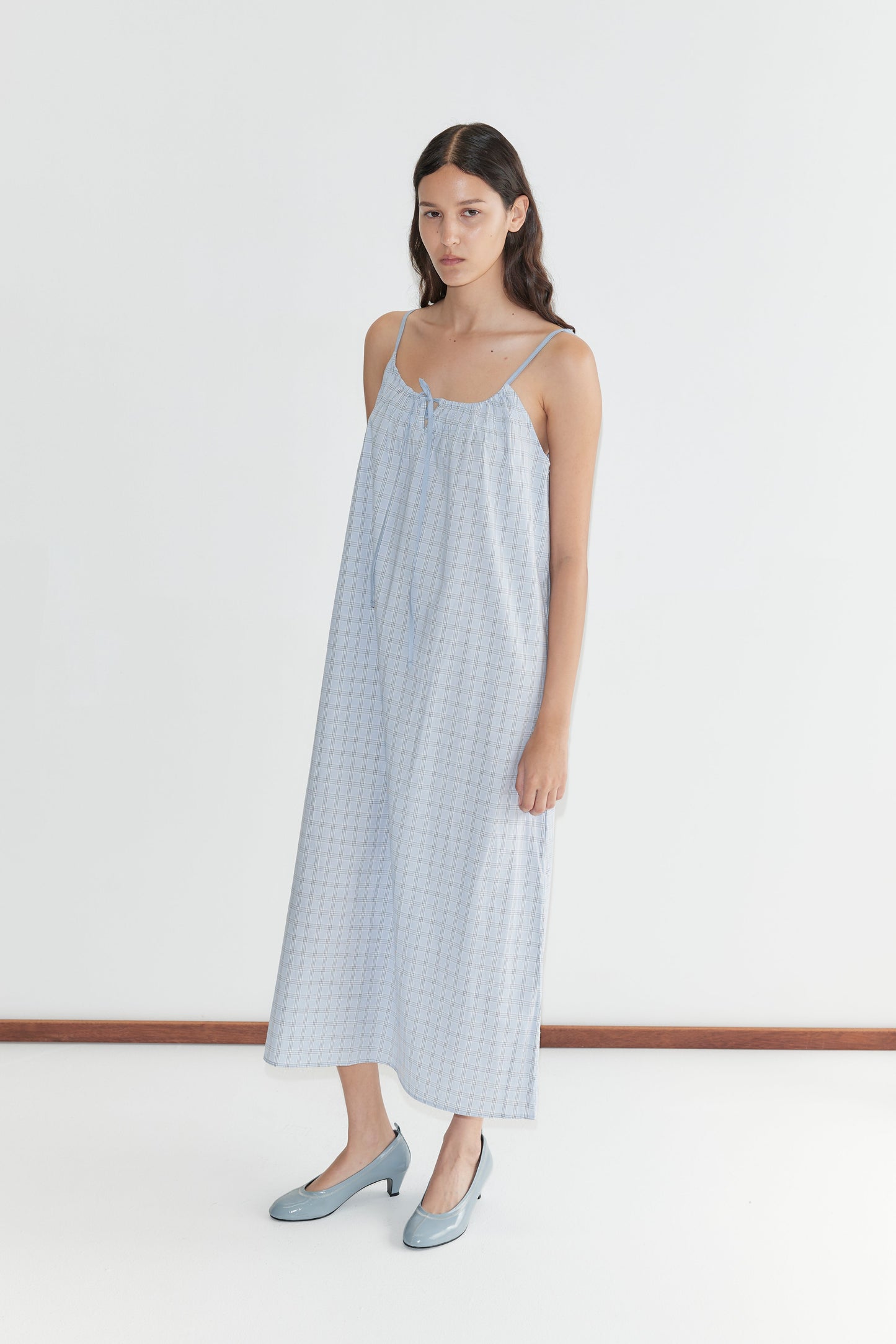 A woman with long dark hair stands against a white background wearing the Light Maxi Dress - Powder Check by Deiji Studios, a loose, sleeveless blue checkered organic cotton maxi dress. She pairs it with blue heels and maintains a neutral expression.