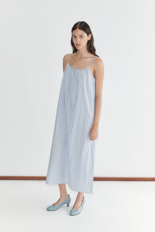 A woman with long dark hair stands against a white background wearing the Light Maxi Dress - Powder Check by Deiji Studios, a loose, sleeveless blue checkered organic cotton maxi dress. She pairs it with blue heels and maintains a neutral expression.