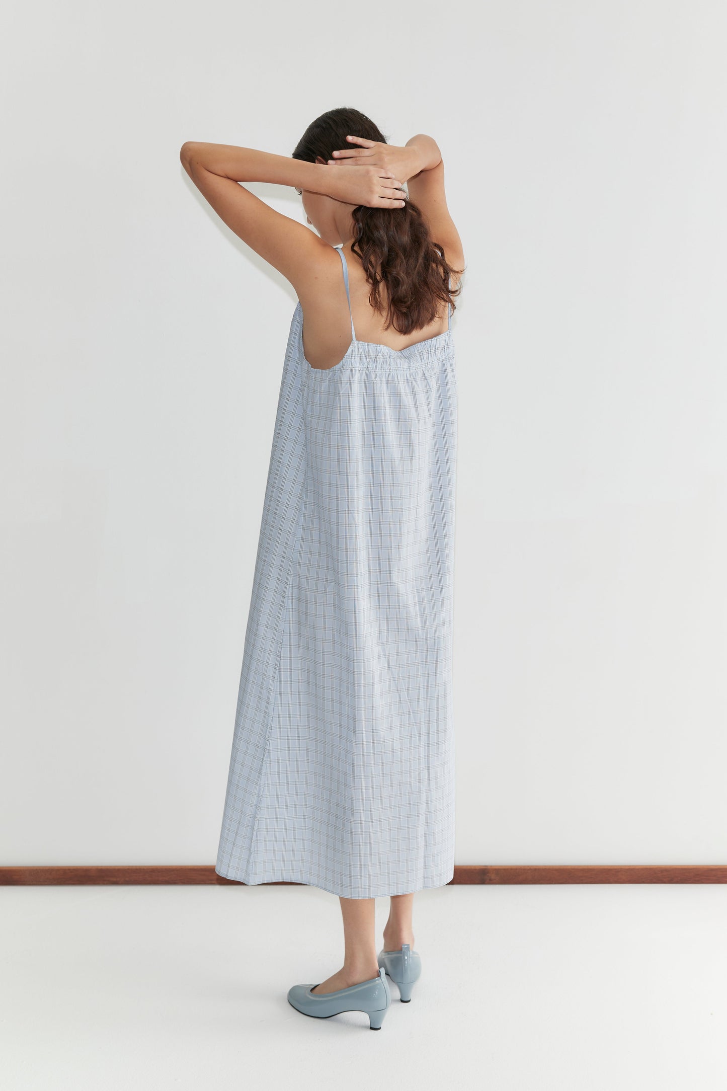 A woman in the Light Maxi Dress - Powder Check from Deiji Studios and blue shoes stands with her back to the camera, arms raised and hands resting on her head, facing a white wall.