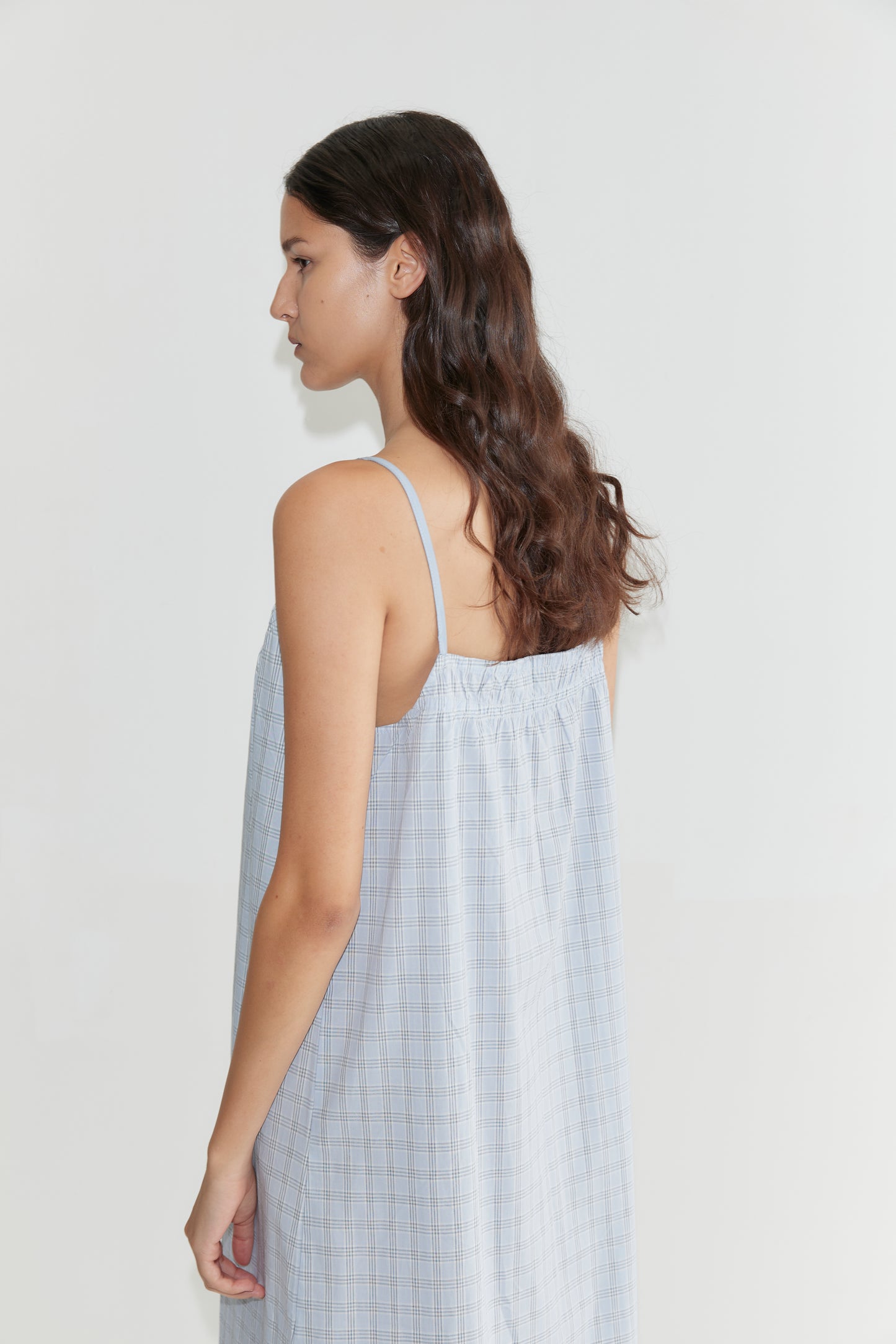 A woman with long, wavy brown hair wears the Light Maxi Dress - Powder Check, a loose maxi dress with thin straps, standing sideways against a plain white background.
