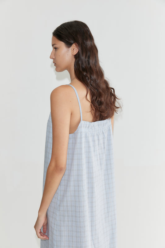 A woman with long, wavy brown hair wears the Light Maxi Dress - Powder Check, a loose maxi dress with thin straps, standing sideways against a plain white background.