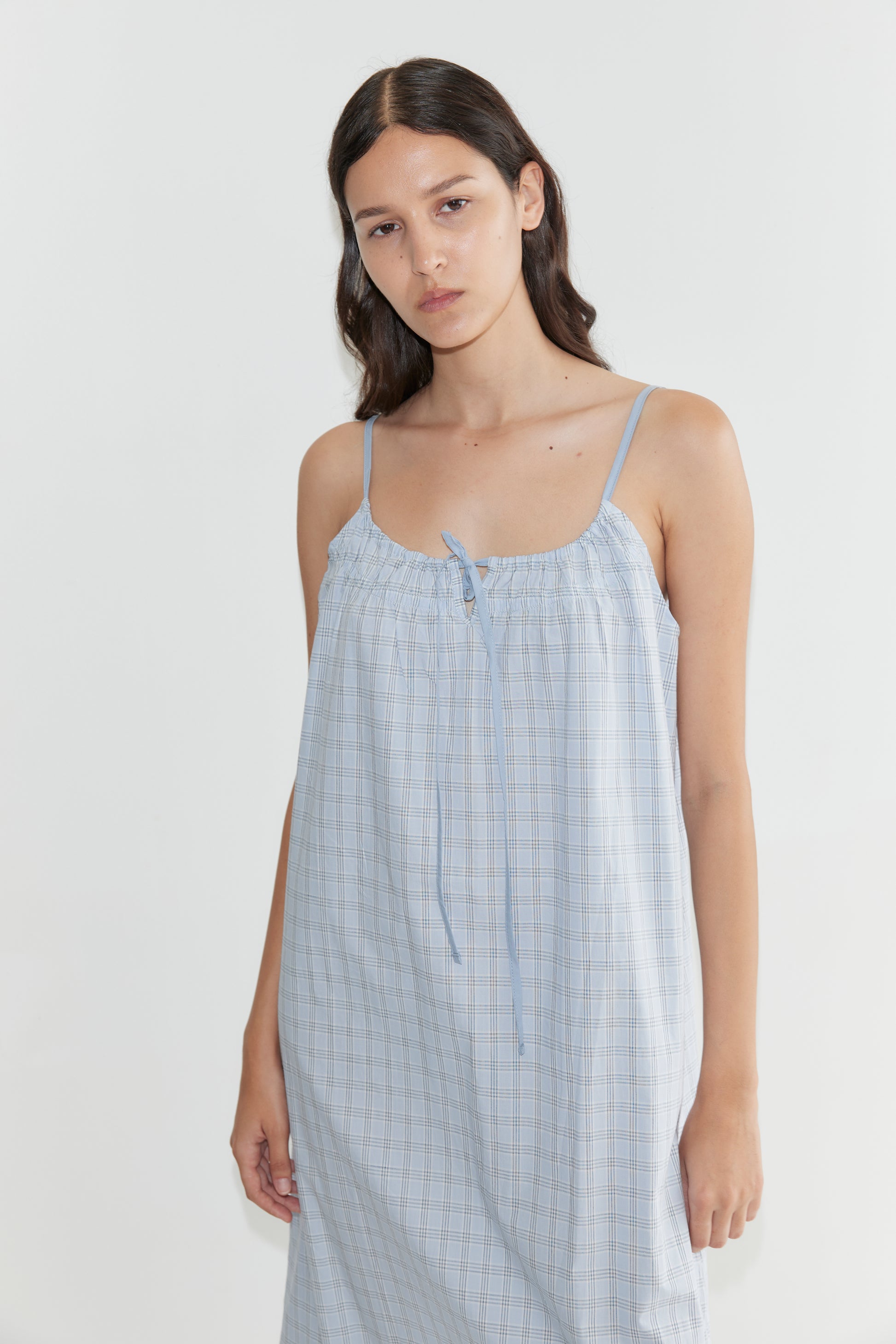A woman with long dark hair wears the Light Maxi Dress - Powder Check by Deiji Studios, a light blue, sleeveless organic cotton maxi with thin straps and tie neckline, featuring a grid pattern. She stands against a plain white background.