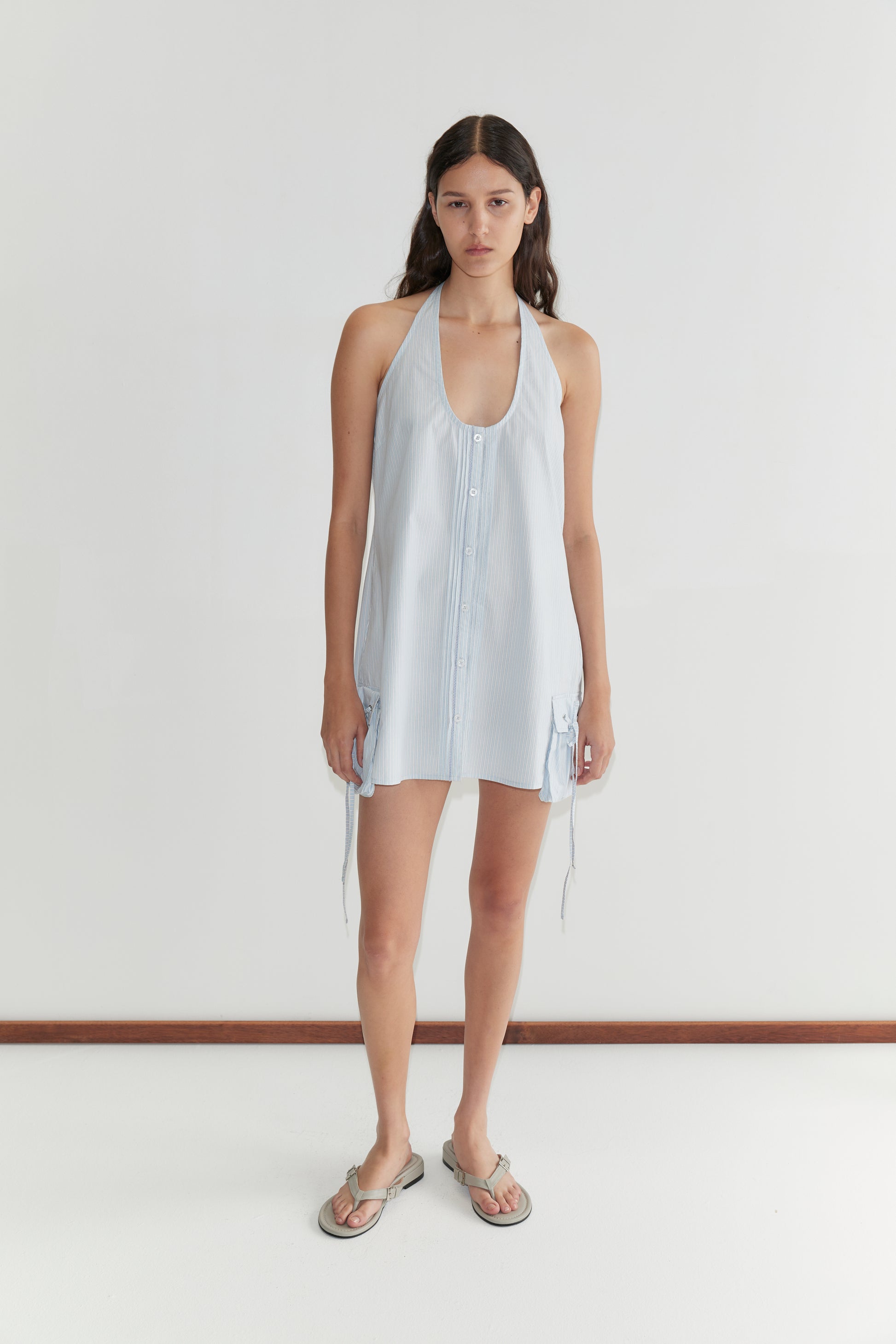 A woman with long brown hair stands against a white background, wearing the Pocket Halter Dress - Mist Stripe by Deiji Studios and flat sandals. She has a neutral expression with her arms at her sides.