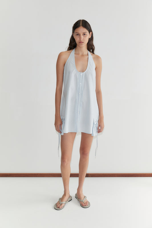 A woman with long brown hair stands against a white background, wearing the Pocket Halter Dress - Mist Stripe by Deiji Studios and flat sandals. She has a neutral expression with her arms at her sides.