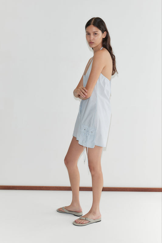 A woman with long dark hair stands sideways against a white wall, wearing the Pocket Halter Dress - Mist Stripe from Deiji Studios and gray flip-flops. Her arms are relaxed by her sides and she has a neutral expression.