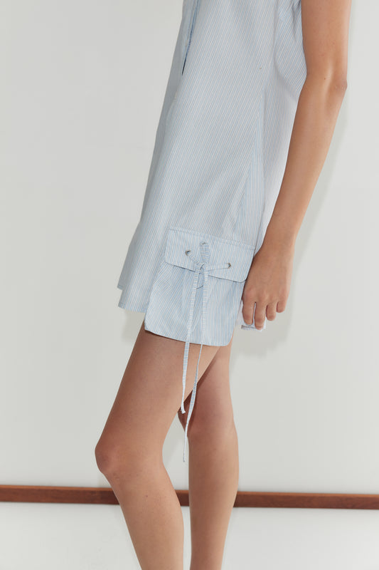 A person wears the Pocket Halter Dress - Mist Stripe by Deiji Studios, a light blue pinstriped adjustable halter dress with a flap side pocket and dangling tie detail, standing against a plain white background.