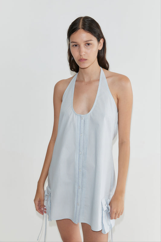 A woman with long dark hair wears the Pocket Halter Dress - Mist Stripe, a light blue, sleeveless dress with a deep neckline and tie details at the hem, standing against a plain white background.