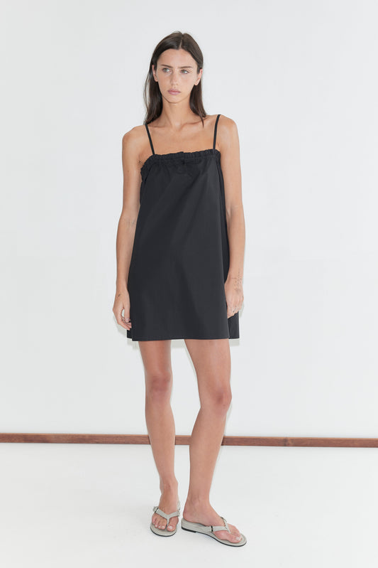 A woman with long dark hair stands against a plain white background wearing the Frill Placket Dress - Black by Deiji Studios, a black A-line mini dress with thin straps and a frilled placket neckline, with grey flip-flops.