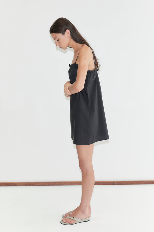 A woman with long dark hair stands in profile against a plain white background, looking down, wearing the Frill Placket Dress - Black by Deiji Studios, a black A-line mini dress with thin straps, and flip-flops.