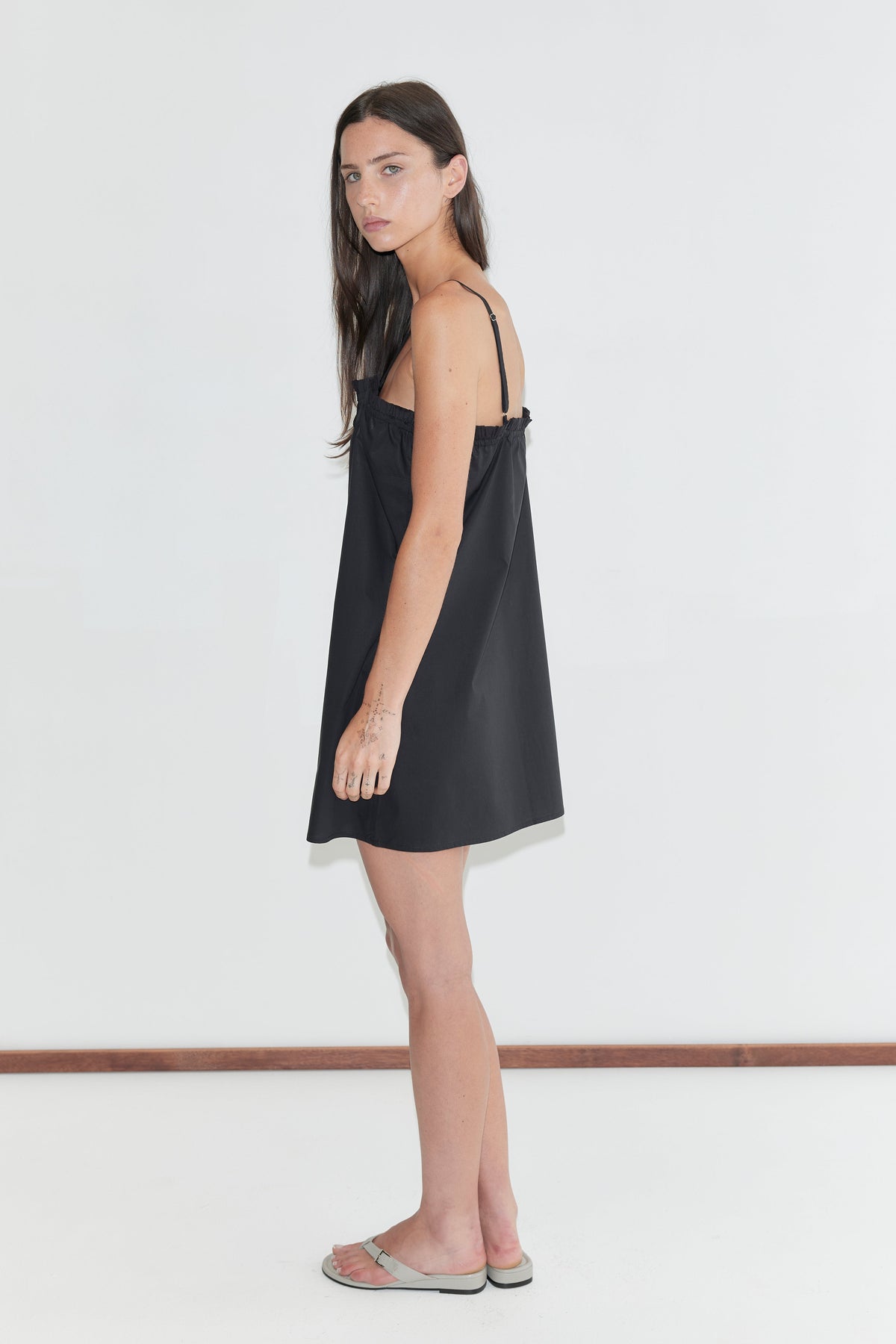 A woman with long dark hair stands in profile against a plain white background wearing the Frill Placket Dress - Black by Deiji Studios, a black A-line mini dress with thin straps, and flip-flops.