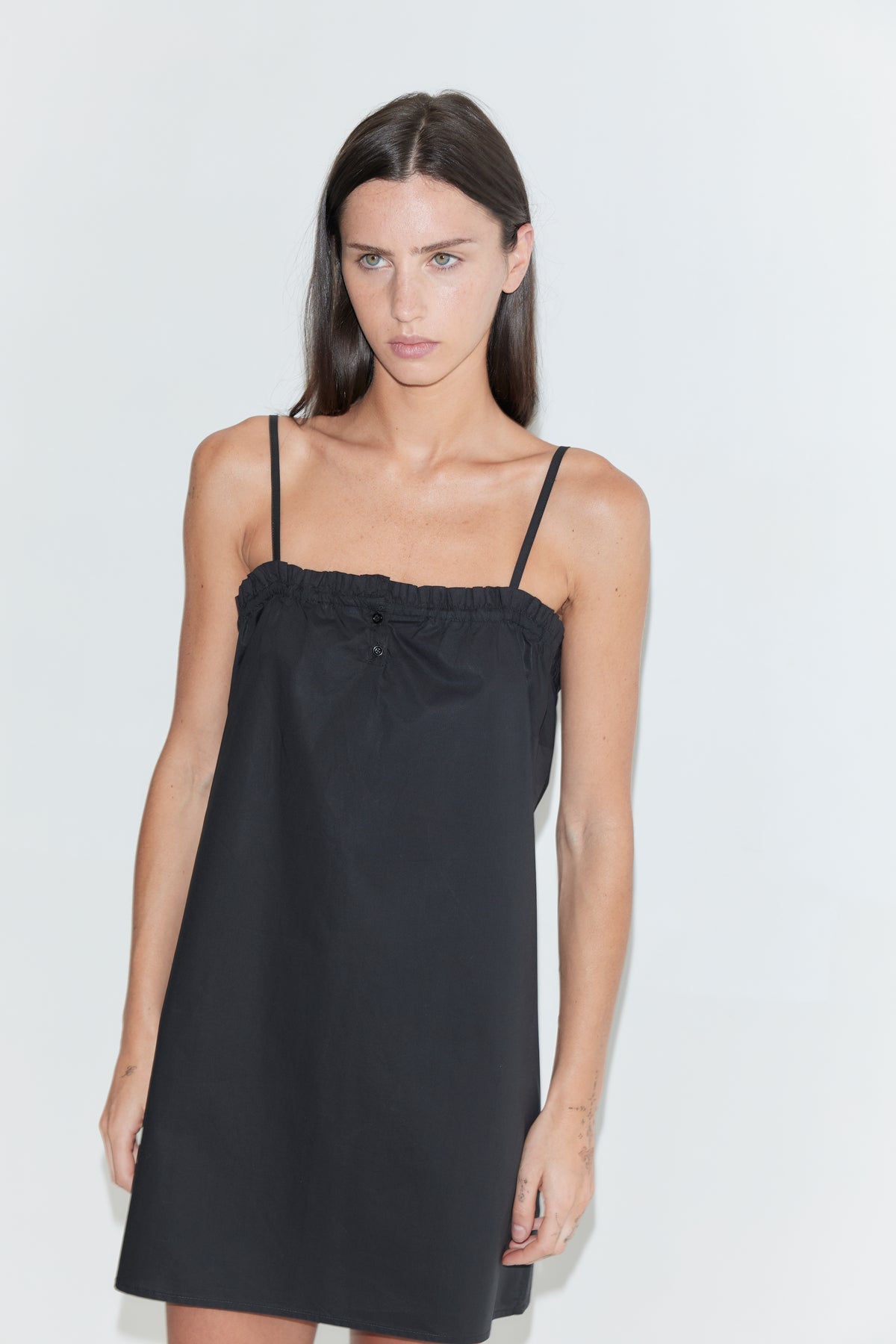 A woman with long dark hair stands against a plain white background wearing the Frill Placket Dress - Black by Deiji Studios, a black mini dress with thin straps and a gathered, buttoned frill placket at the neckline.