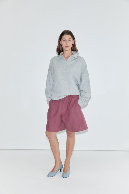 A person wearing a light gray sweatshirt, the "Half Short - Brick" linen shorts with an elasticated waistband and wide leg opening in a knee-length style, and blue heels stands against a plain white background, facing the camera with hands in their pockets.