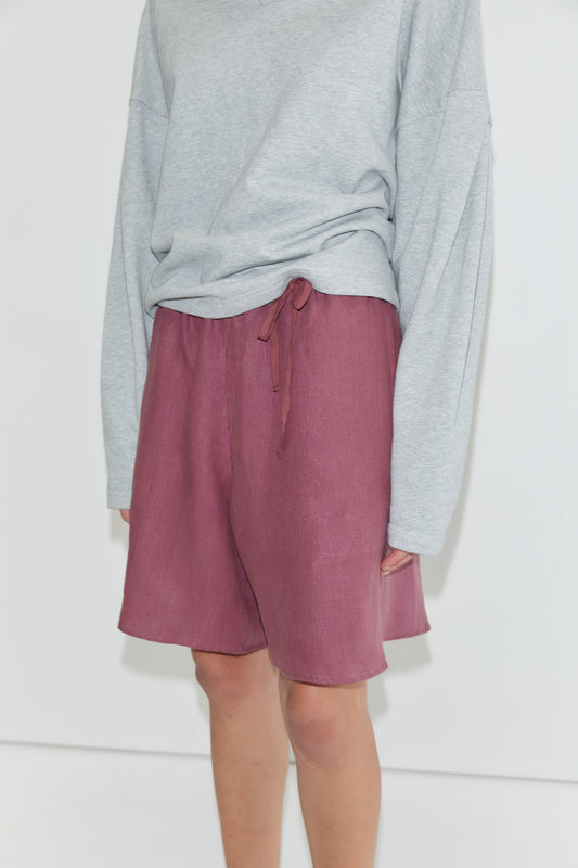 A person wearing a loose grey long-sleeve shirt and high-waisted, knee-length Half Short - Brick relaxed linen shorts with an elasticated waistband and wide leg opening, standing against a plain white background.