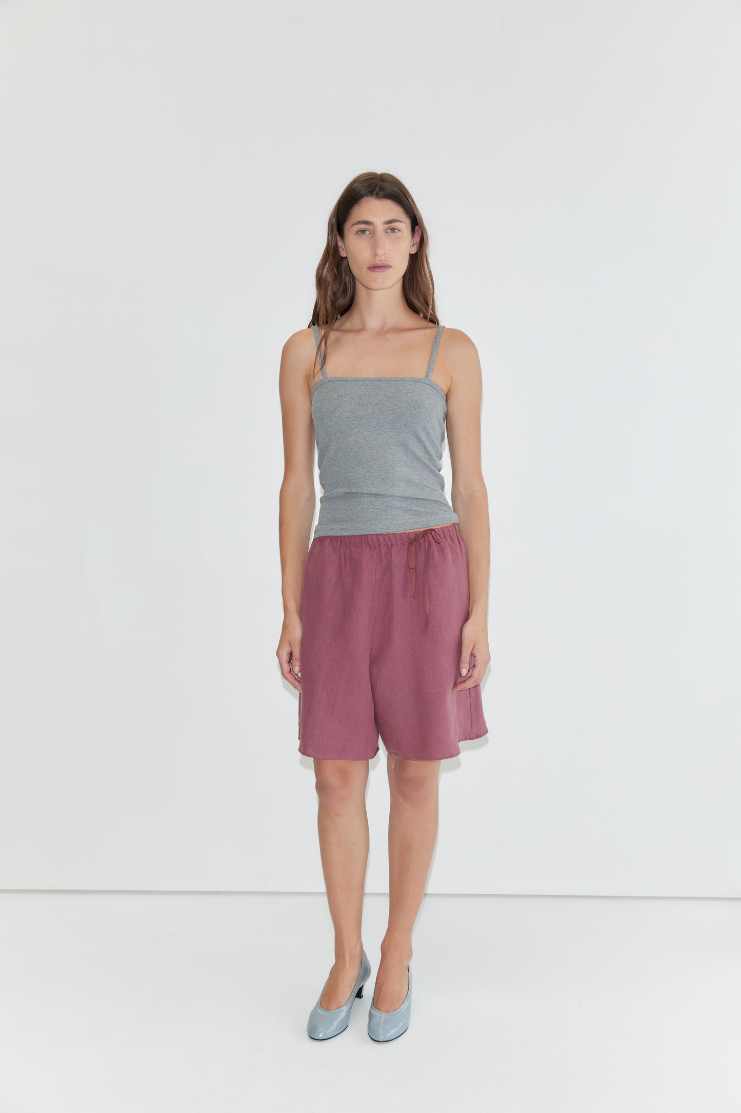 A person stands against a plain white background, wearing a gray spaghetti strap top, Half Short - Brick with an elasticated waistband and wide leg opening in maroon, paired with light blue shoes.