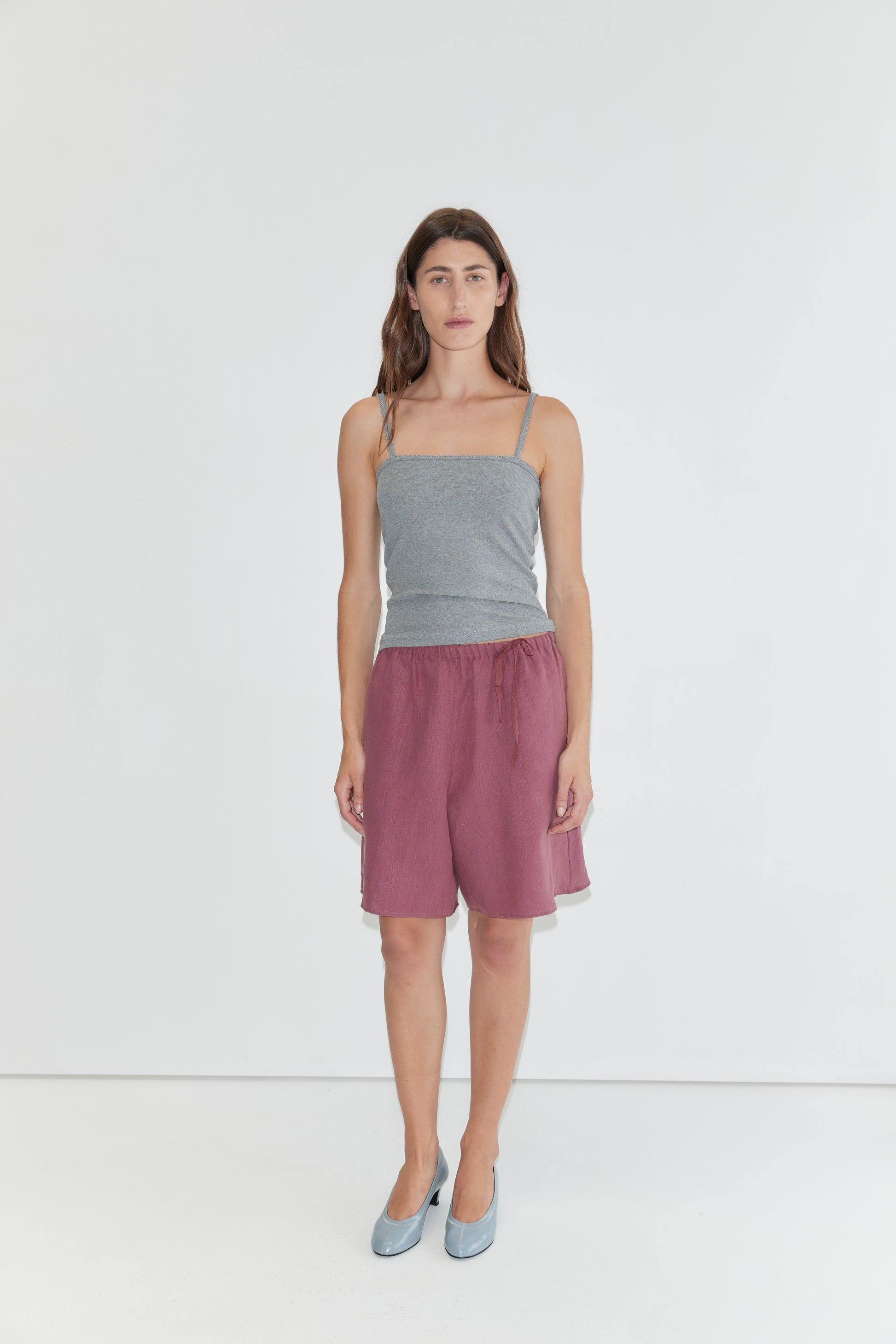 A person stands against a plain white background, wearing a gray spaghetti strap top, Half Short - Brick with an elasticated waistband and wide leg opening in maroon, paired with light blue shoes.