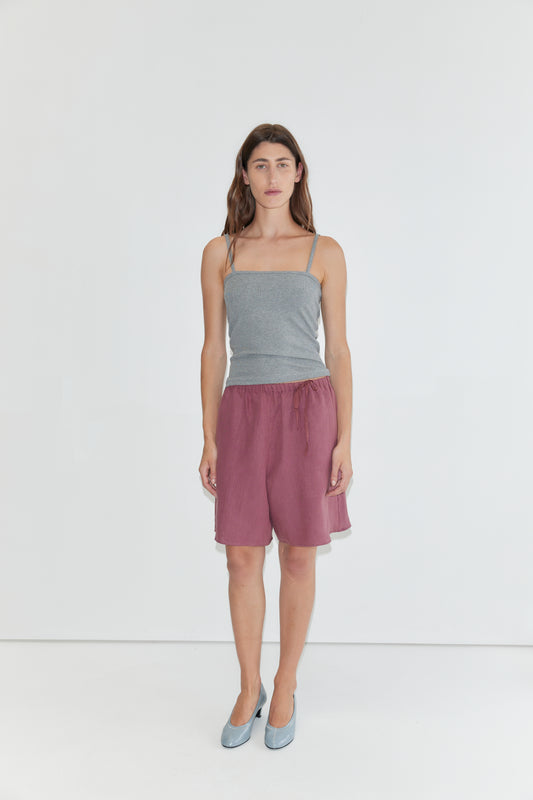 A person stands against a plain white background, wearing a gray spaghetti strap top, Half Short - Brick with an elasticated waistband and wide leg opening in maroon, paired with light blue shoes.
