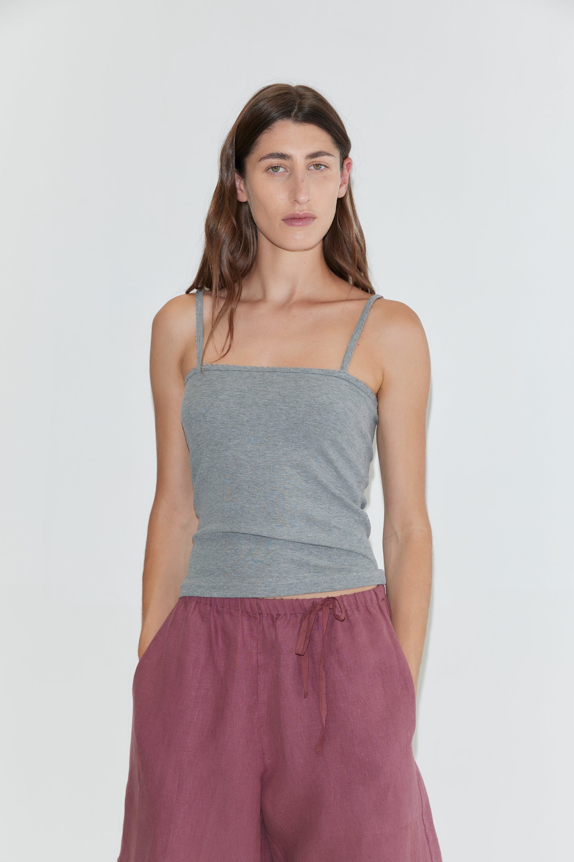 A woman with long brown hair, dressed in a gray spaghetti strap top and the Half Short - Brick, which features a wide leg opening and an elasticated waistband, stands against a plain white background with her hands in her pockets.