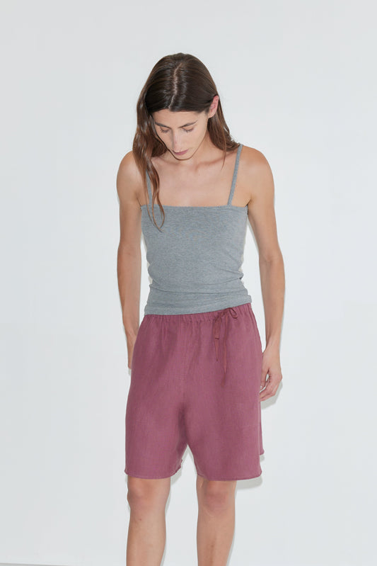 A person with long brown hair, wearing the Scallop Tank - Grey and loose maroon shorts, looks down while standing against a plain white background.