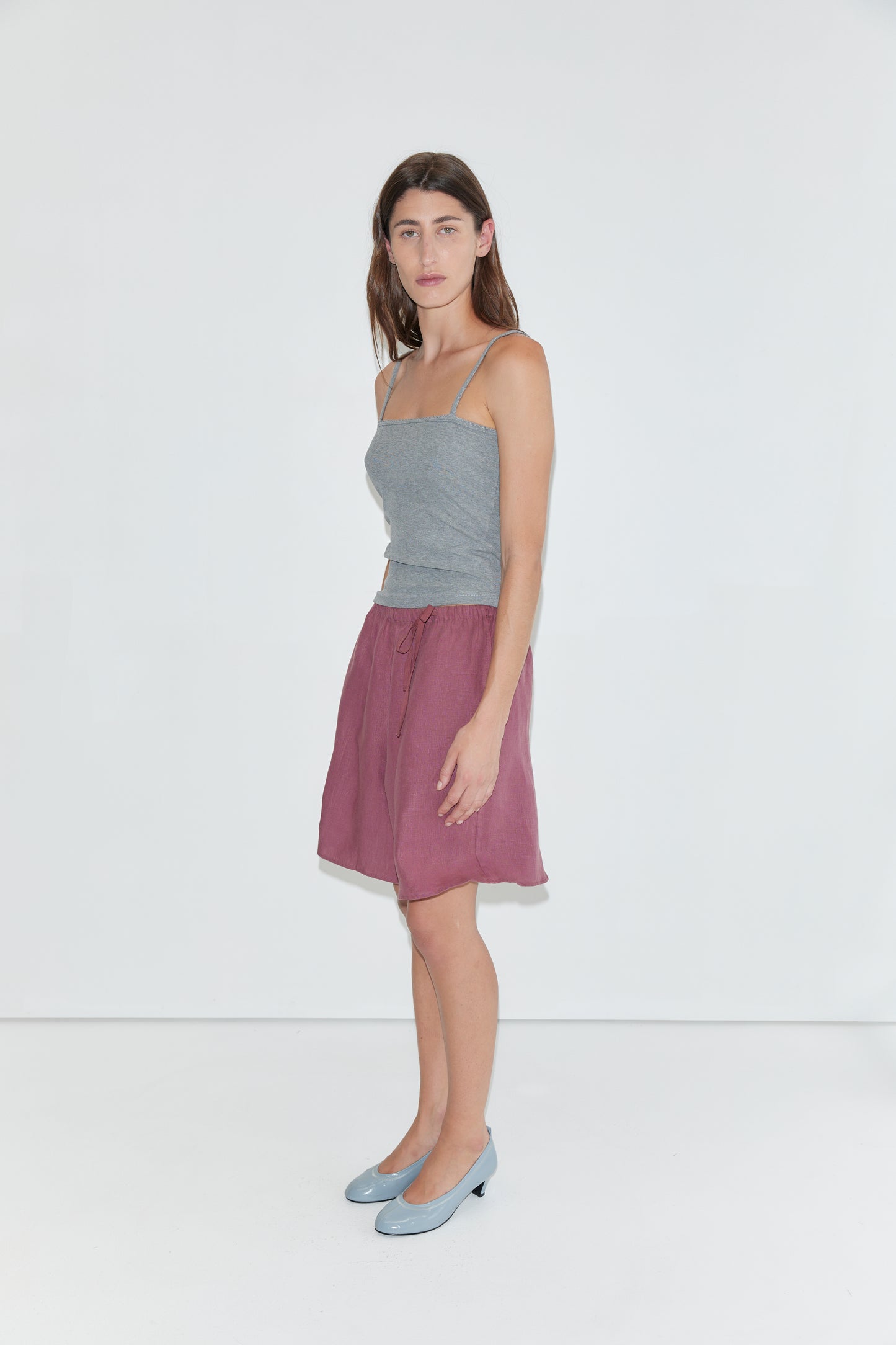 A woman stands against a plain white background, wearing the Scallop Tank - Grey with spaghetti straps, paired with a mauve knee-length skirt and light blue low-heeled shoes.