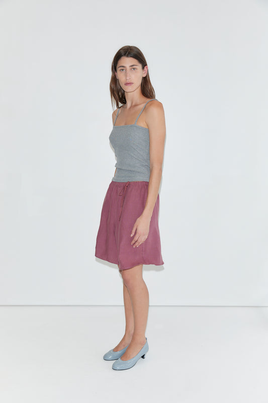 A woman stands against a plain white background, wearing the Scallop Tank - Grey with spaghetti straps, paired with a mauve knee-length skirt and light blue low-heeled shoes.