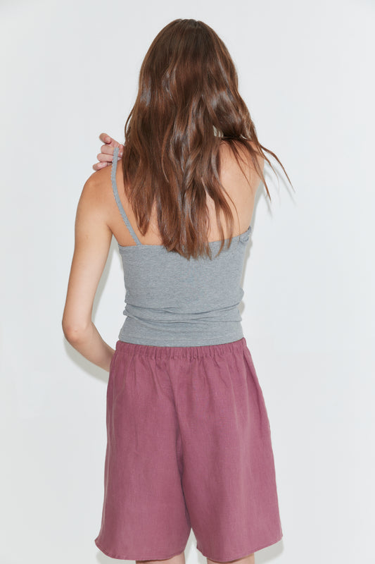 Wearing a gray tank top and the Half Short - Brick, which features an elasticated waistband and wide leg opening, a person with long brown hair stands facing away from the camera against a plain white background.