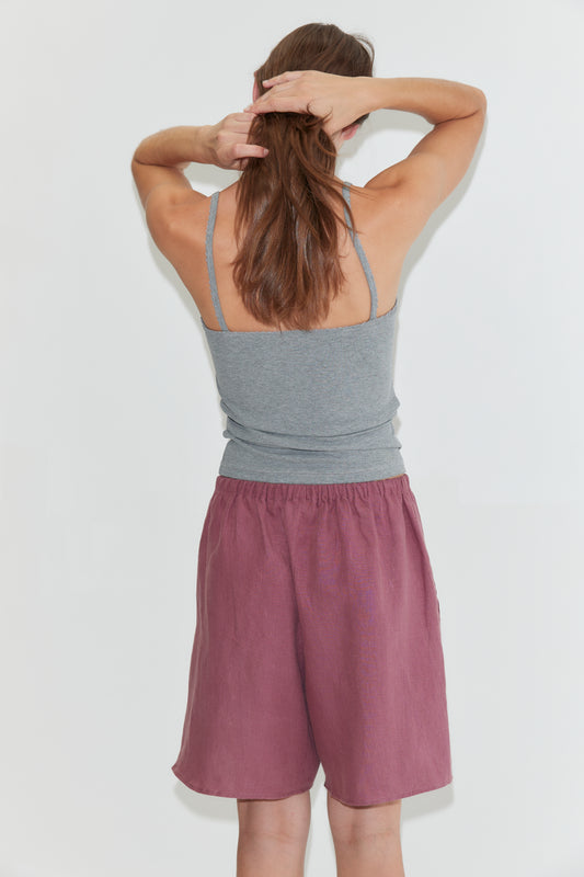Wearing the Scallop Tank in Grey, a person with long brown hair, paired with mauve shorts, stands facing away and ties their hair with both hands against a plain white background.