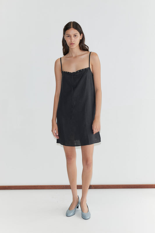 A woman with long brown hair and a neutral expression stands against a white background, wearing the Weave Mini - Black dress from Deiji Studios with adjustable straps and light blue flat shoes.