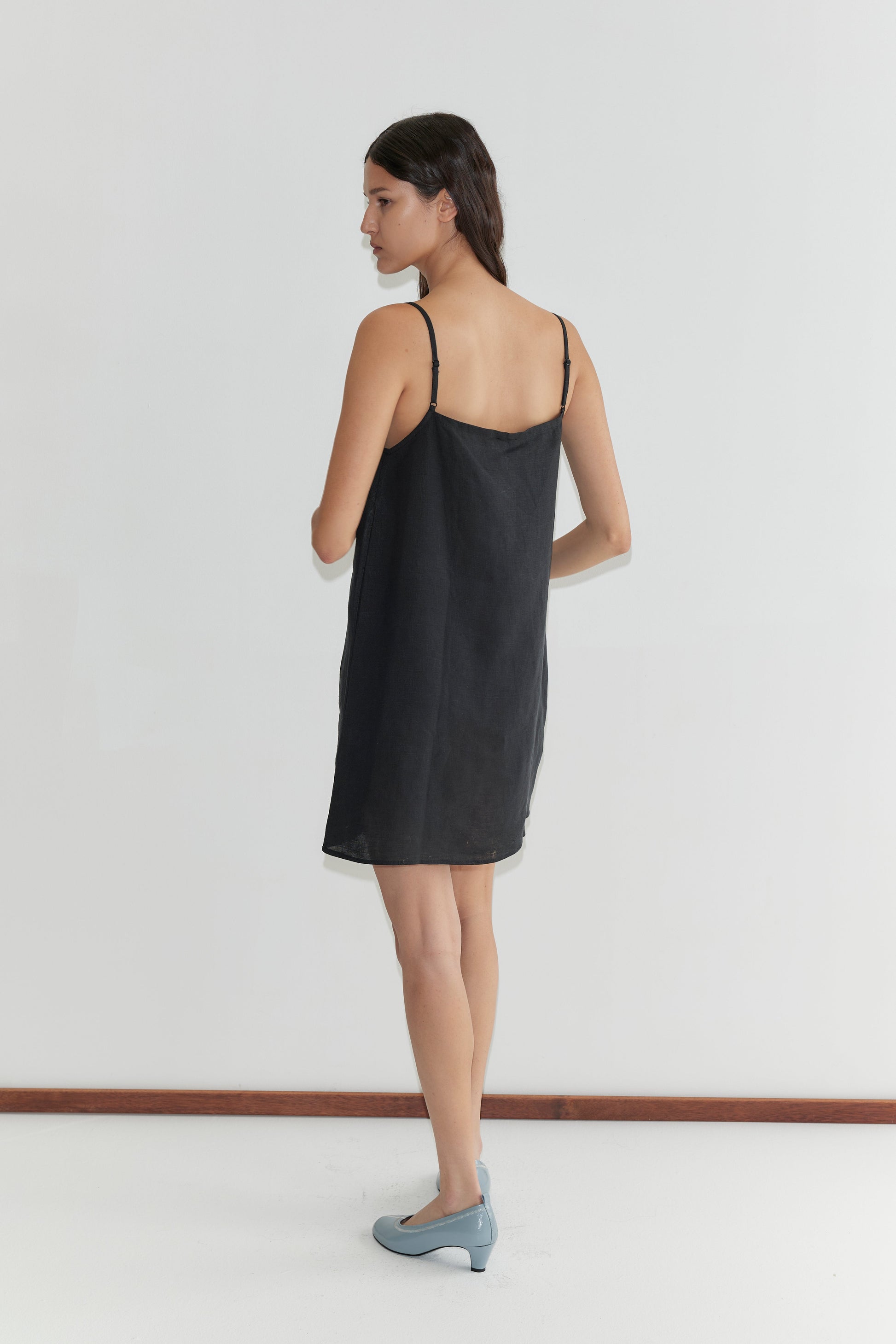 A woman with long dark hair wears the Weave Mini - Black, a simple black linen mini dress with adjustable straps, paired with light blue shoes. She stands indoors against a plain white wall, facing slightly to the side.